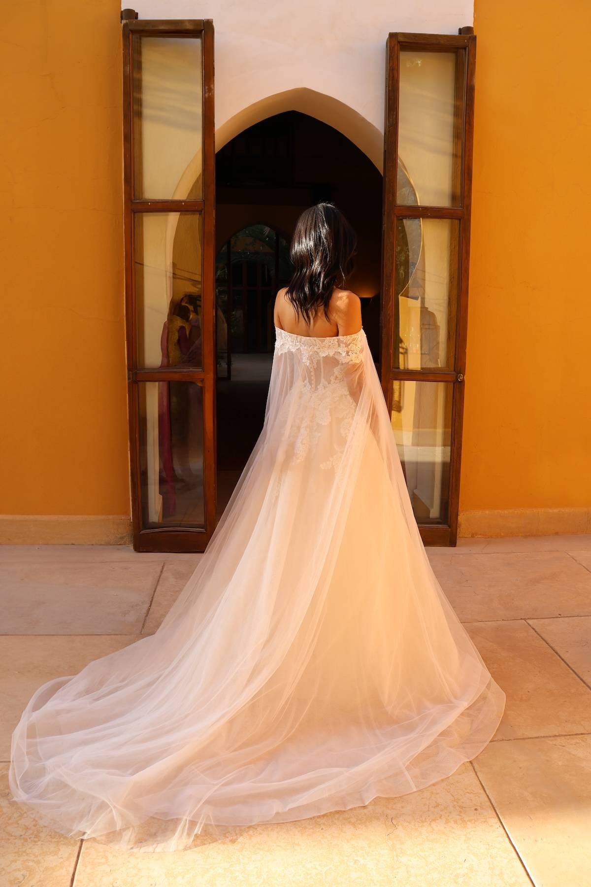 Robe de mariée Rosée d’Or en dentelle avec cape longue posée sur les épaules, descendant sur la traîne.