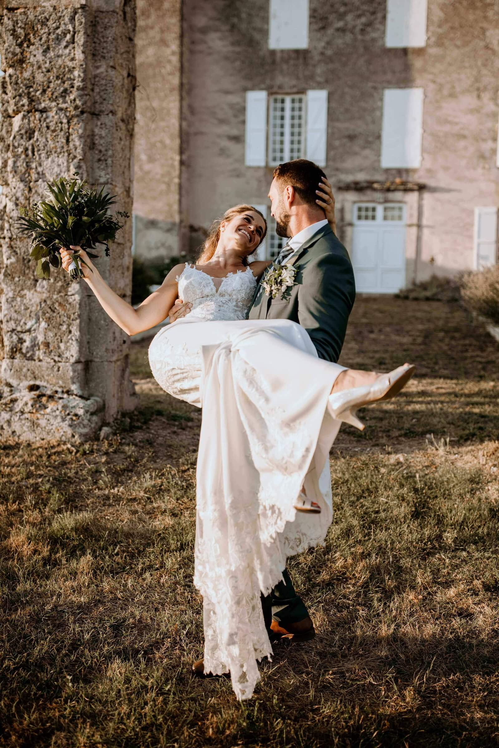 Julien porte Sylvie dans ses bras près d’un arbre, la jupe de sa robe de mariée déployé